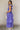 Full body side view of model wearing the Harlow Blue & Purple Floral Ruffle Midi Dress which features blue and purple sheer fabric, floral print, light purple metallic, blue lining, three tiered skirt, midi length, plunge neckline, adjustable ruffle strap