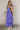 Full body side view of model wearing the Harlow Blue & Purple Floral Ruffle Midi Dress which features blue and purple sheer fabric, floral print, light purple metallic, blue lining, three tiered skirt, midi length, plunge neckline, adjustable ruffle strap