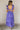 Full body back view of model wearing the Harlow Blue & Purple Floral Ruffle Midi Dress which features blue and purple sheer fabric, floral print, light purple metallic, blue lining, three tiered skirt, midi length, plunge neckline, adjustable ruffle strap