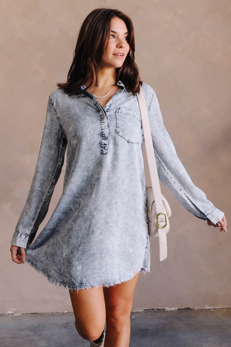 Front view of model wearing the Talk The Talk Dress In Blue that has washed blue denim fabric, mini length, a frayed hem, a hidden quarter button up, a collar, a chest pocket, and long sleeves.