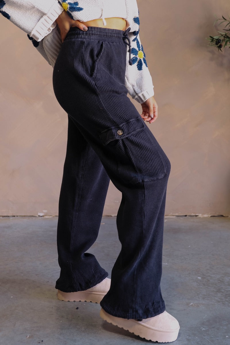 Side view of model wearing the Ayla Washed Black Cargo Adjustable Jogger Pants which features washed black ribbed fabric, two front pockets, elastic waistband with drawstring tie, two side cargo pockets with tortoise button and wide pant legs with drawstr