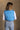 Back view of model wearing the Sutton Blue Cropped Puffer Vest that has blue puffer fabric, a  3 tiered design, medium blue lining, a cropped waist,pockets, a front zipper, a drawstring, a high neck and a sleeveless design.