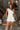Front view of model wearing the All My Love Ruffle Romper which features white fabric, pockets on each side, a tie around the waist, one shoulder ruffle strap, and a monochromatic side zipper with a hook closure.
