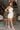 Full body side view of model wearing the All My Love Ruffle Romper which features white fabric, pockets on each side, a tie around the waist, one shoulder ruffle strap, and a monochromatic side zipper with a hook closure.