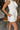 Close up side view of model wearing the All My Love Ruffle Romper which features white fabric, pockets on each side, a tie around the waist, one shoulder ruffle strap, and a monochromatic side zipper with a hook closure.