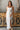 Full body view of model wearing the Falling For You Midi Dress which features white sheer fabric with embroidered white floral print, white skirt lining, tan upper lining, a midi length skirt with a side slit, boning throughout, a sweetheart neckline, adj