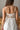 Back view of model wearing the Falling For You Midi Dress which features white sheer fabric with embroidered white floral print, white skirt lining, tan upper lining, a midi length skirt with a side slit, boning throughout, a sweetheart neckline, adjustab