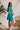 Full side view of model wearing the All For You Dress that has teal fabric with a lace floral pattern, mini length, teal lining, a tie around the waist, a surplice neckline, and short puff sleeves with elastic trim