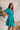Side view of model wearing the All For You Dress that has teal fabric with a lace floral pattern, mini length, teal lining, a tie around the waist, a surplice neckline, and short puff sleeves with elastic trim