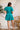 Full back view of model wearing the All For You Dress that has teal fabric with a lace floral pattern, mini length, teal lining, a tie around the waist, a surplice neckline, and short puff sleeves with elastic trim