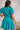 Back view of model wearing the All For You Dress that has teal fabric with a lace floral pattern, mini length, teal lining, a tie around the waist, a surplice neckline, and short puff sleeves with elastic trim