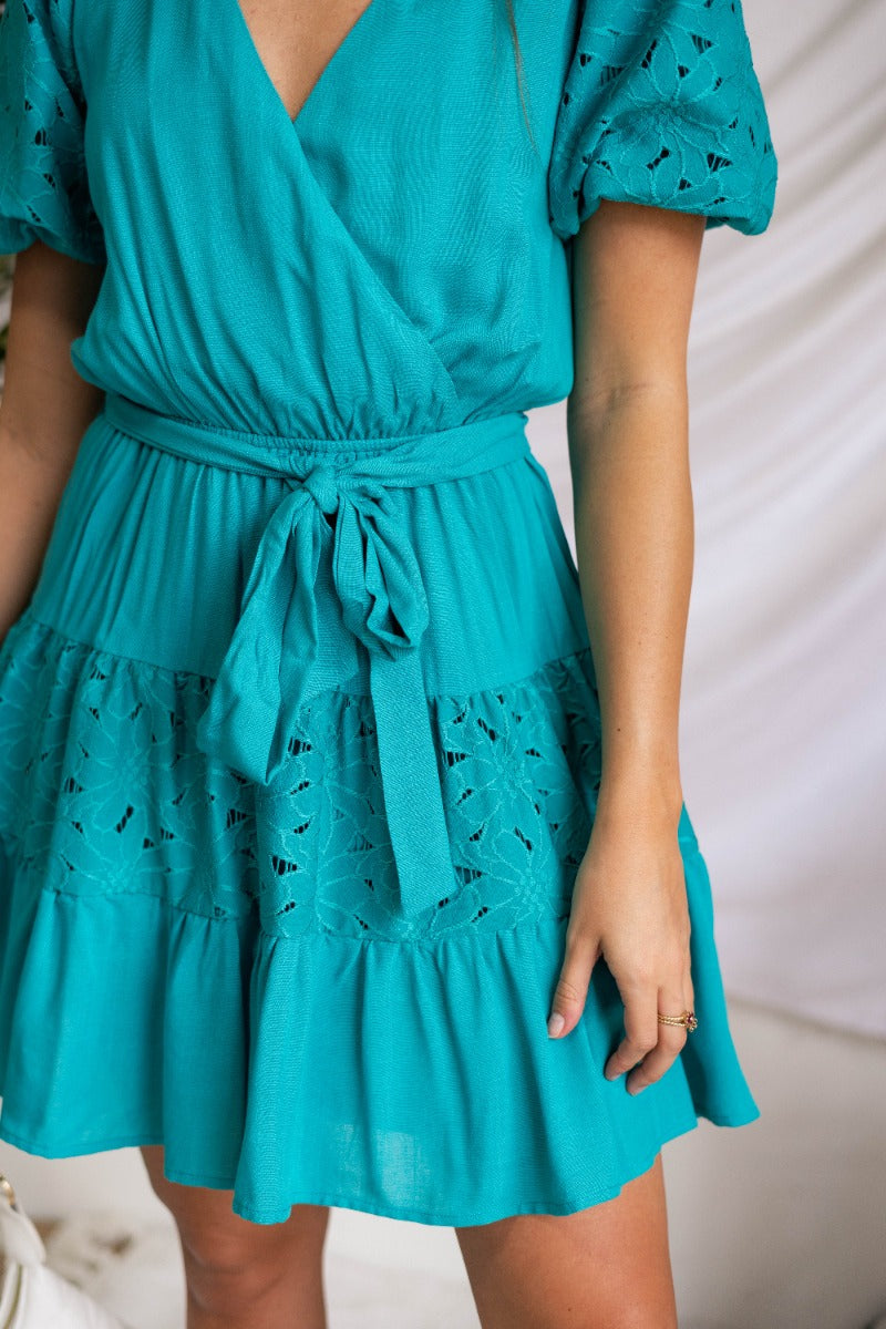 Close lower front view of model wearing the All For You Dress that has teal fabric with a lace floral pattern, mini length, teal lining, a tie around the waist, a surplice neckline, and short puff sleeves with elastic trim