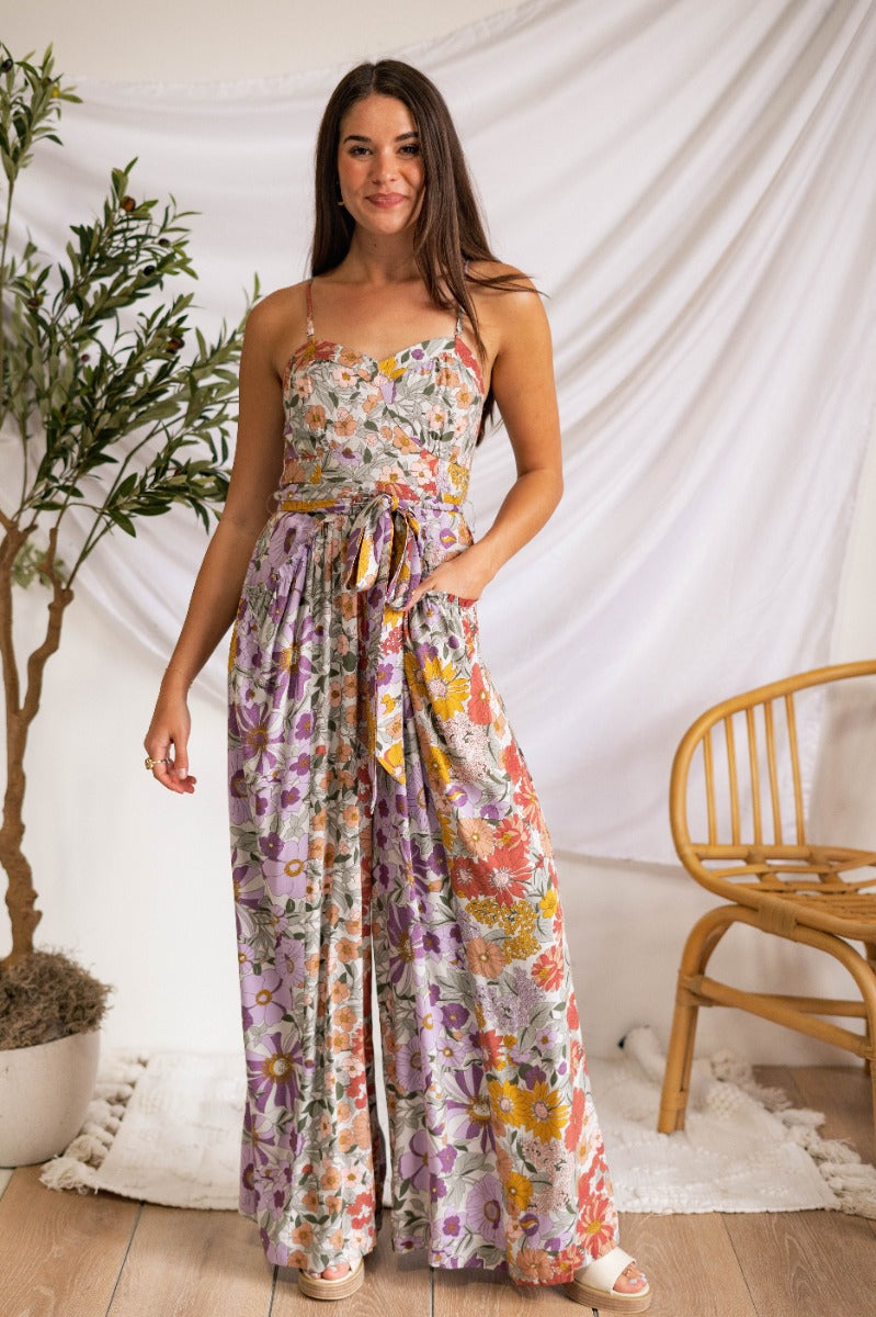 Full body view of model wearing the Send Flowers Jumpsuit which features lavender fabric with a green, mustard, peach, light pink and red floral print, flared pant legs, ruffle details, two side pockets, a sweetheart neckline, adjustable straps, a smocked