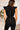 Upper back view of model wearing the She's All That Denim Jumpsuit that has black denim fabric, cutout details, a surplice neck with ruffles, a sleeveless design, flare pant legs, and a back zipper.