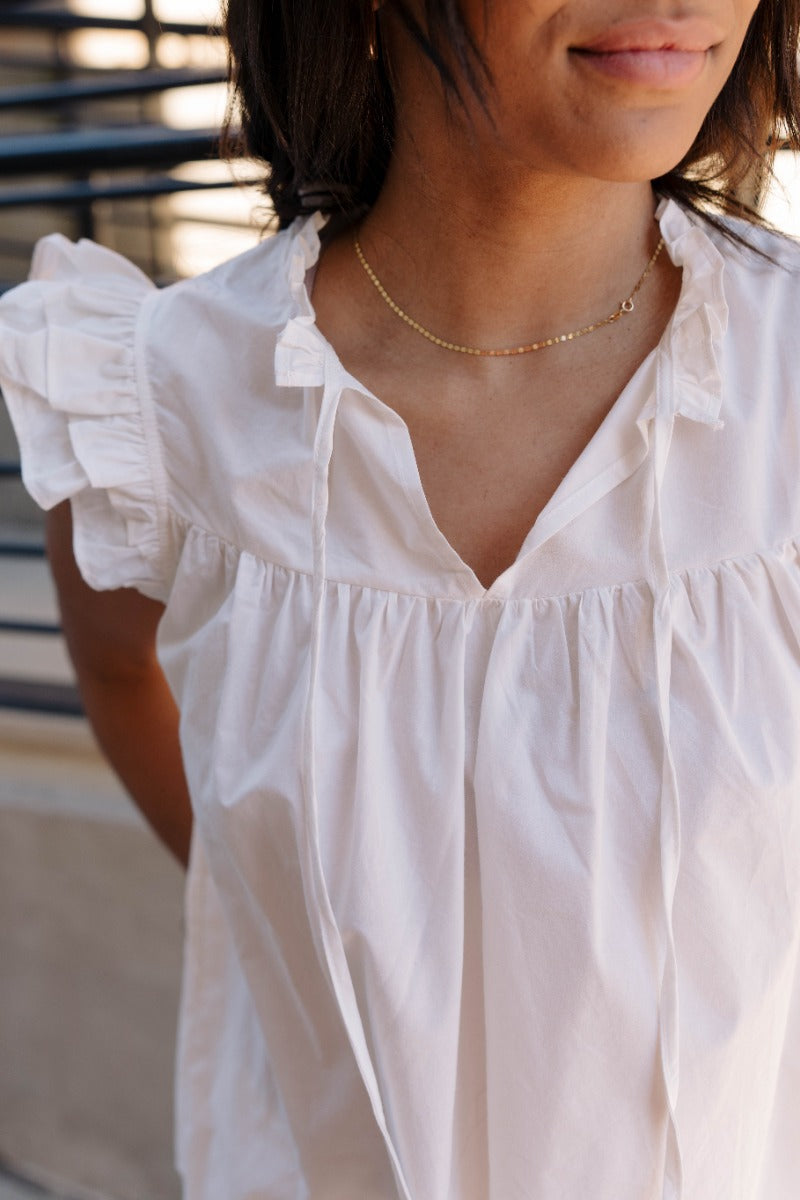 Close front view of model wearing the Emory White Sleeveless Ruffle Top that features off white cotton fabric, a ruffled notched neckline with ties, and a sleeveless body with ruffle details.