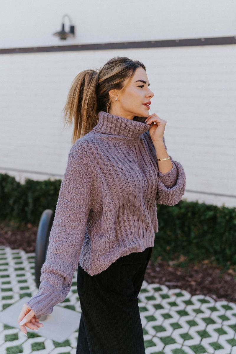 Side view of the Days Go By Sweater in Grey that features grey cable knit fabric on the front, loose turtleneck, popcorn detailing on the back and sleeves and long sleeves with cable knit wrists.