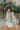 Side view of model wearing the Forever Yours Top, which has a sage green fabric , a round neckline, long sleeves, a back tie closure at the neck, and a flowy fit