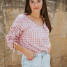 Frontal view of the Cruel Summer Top that features a pink colored material, a white gingham print, a V neckline, a long ruched sleeve with adjustable ties, and a flowy fit.