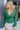 Front view of model wearing the Follow Your Heart Top, which features a green colored material, a V surplice neckline, a long sleeve, a knot detail at the waist, and a tiered ruffle bottom