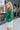 Side view of model wearing the Follow Your Heart Top, which features a green colored material, a V surplice neckline, a long sleeve, a knot detail at the waist, and a tiered ruffle bottom