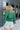 Back view of model wearing the Follow Your Heart Top, which features a green colored material, a V surplice neckline, a long sleeve, a knot detail at the waist, and a tiered ruffle bottom
