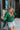 Front view of model wearing the Follow Your Heart Top, which features a green colored material, a V surplice neckline, a long sleeve, a knot detail at the waist, and a tiered ruffle bottom