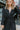 Close-up front view of model wearing the Lost In The Moment Dress in Black, which has a black satin material, a collar neck, a surplice V neckline with a clasp closure, a long cuffed sleeve, a knot detail at the waist, and a midi length.