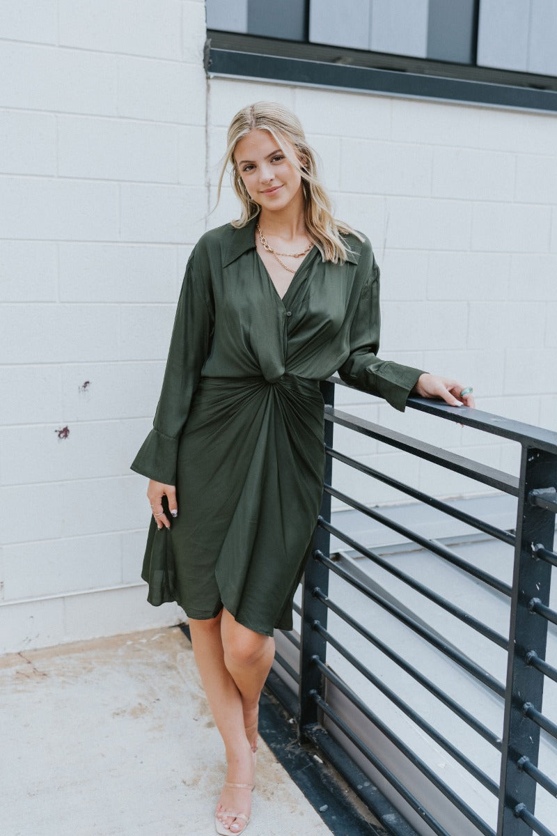 Front view of model wearing the Lost In The Moment Dress in Green, which features a forest green satin material, a collar neck, a surplice V neckline with a clasp closure, long cuffed sleeves, a knot detail at the waist, and a midi length.