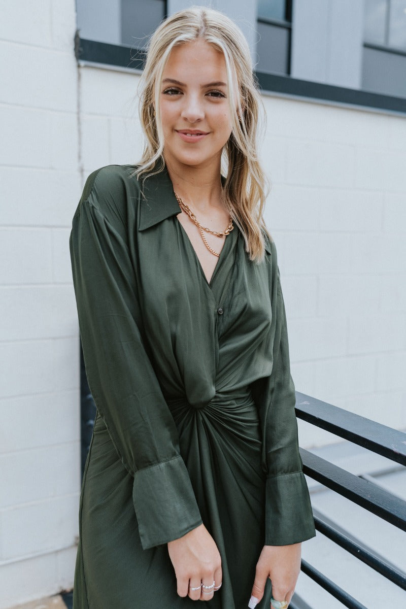 Close-up front view of model wearing the Lost In The Moment Dress in Green, which features a forest green satin material, a collar neck, a surplice V neckline with a clasp closure, long cuffed sleeves, a knot detail at the waist, and a midi length.