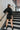 Full body back view of model wearing the Road Trip Romper In Black, which has a collar neck, a button-up front, two front pockets at the chest, long cuffed sleeves, and a tie belt at the waist