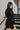 Back view of model wearing the Road Trip Romper In Black, which has a collar neck, a button-up front, two front pockets at the chest, long cuffed sleeves, and a tie belt at the waist
