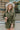 Front view of model wearing the Road Trip Romper In Olive, which has an olive green material, a collar neck, a button-up front, two front pocket, long sleeves and a tie belt at the waist.