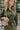 Close-up front view of model wearing the Road Trip Romper In Olive, which has an olive green material, a collar neck, a button-up front, two front pocket, long sleeves and a tie belt at the waist.