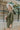 Side view of model wearing the Road Trip Romper In Olive, which has an olive green material, a collar neck, a button-up front, two front pocket, long sleeves and a tie belt at the waist.