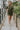 Full body front view of model wearing the Road Trip Romper In Olive, which has an olive green material, a collar neck, a button-up front, two front pocket, long sleeves and a tie belt at the waist.
