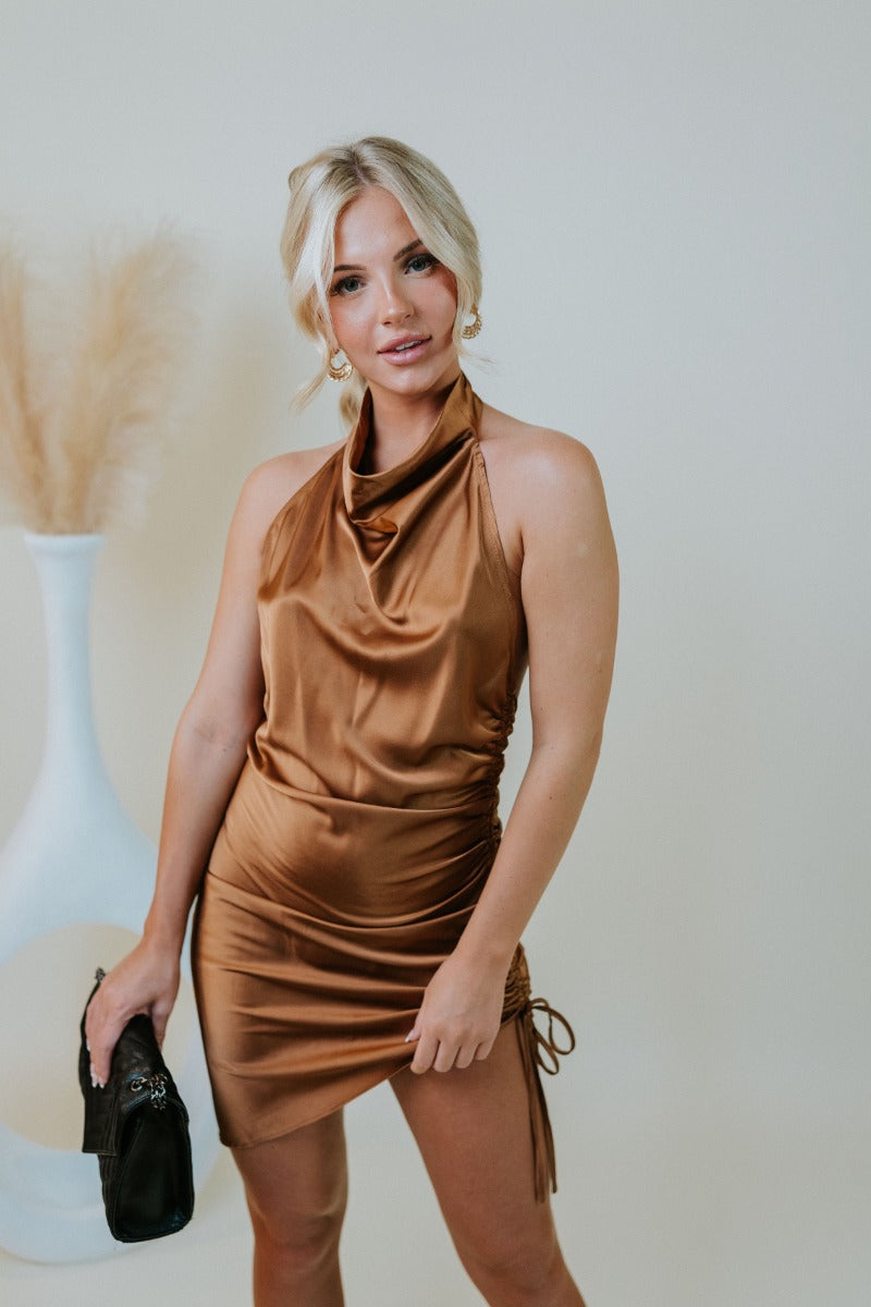 Frontal view of the Wallflower Mini Dress that features a rust silk material, a high cowl neckline, a ruched side with adjustable ties, an asymmetrical hemline, and an open back.