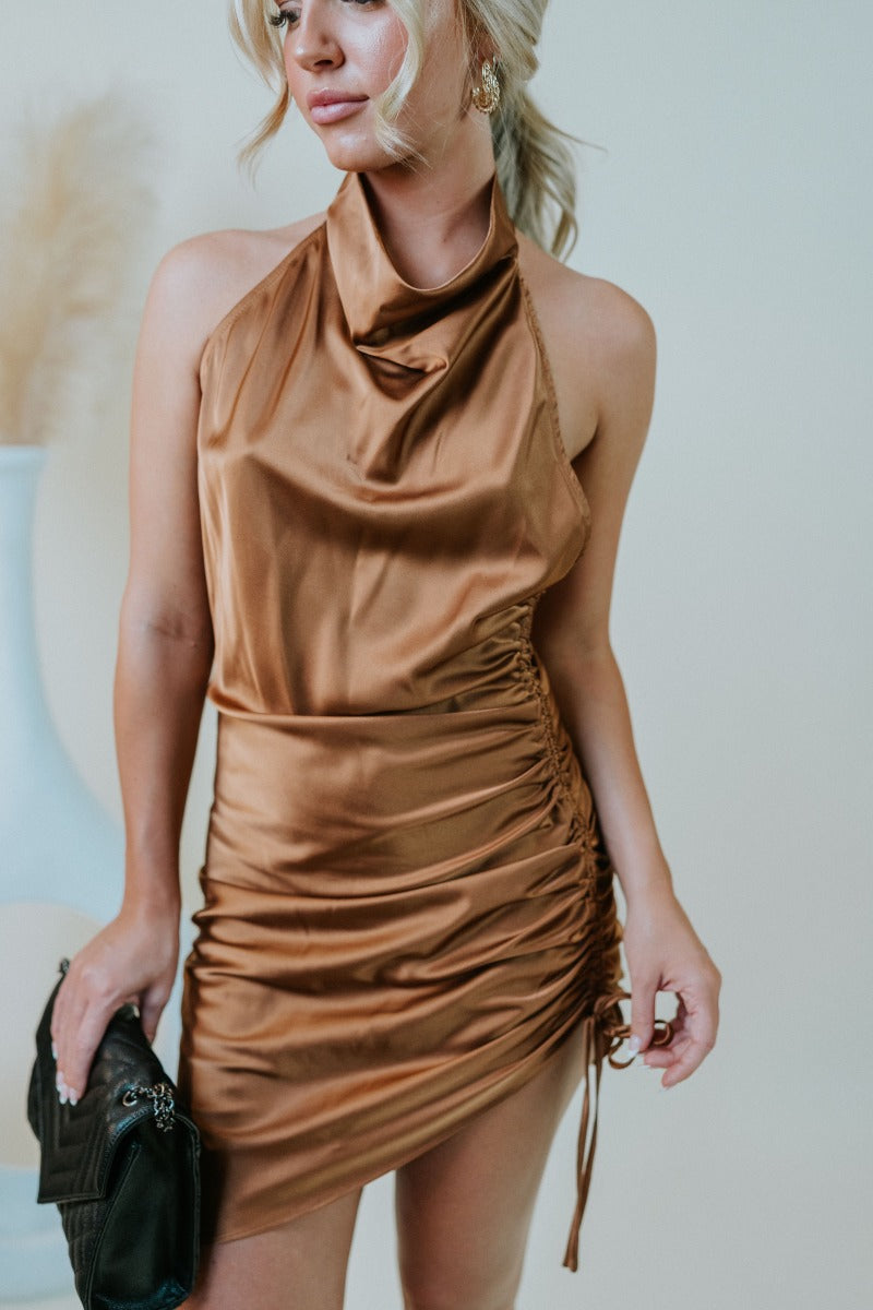 Frontal view of the Wallflower Mini Dress that features a rust silk material, a high cowl neckline, a ruched side with adjustable ties, an asymmetrical hemline, and an open back.