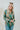 Frontal view of the Leave The Pieces Top that features a green silk material, a multi-colored floral print, a collar neck, a button-up front, long cuffed sleeves, a curved bottom hem, and an oversized fit.