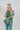 Frontal view of the Leave The Pieces Top that features a green silk material, a multi-colored floral print, a collar neck, a button-up front, long cuffed sleeves, a curved bottom hem, and an oversized fit.