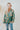 Frontal view of the Leave The Pieces Top that features a green silk material, a multi-colored floral print, a collar neck, a button-up front, long cuffed sleeves, a curved bottom hem, and an oversized fit.
