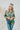 Frontal view of the Leave The Pieces Top that features a green silk material, a multi-colored floral print, a collar neck, a button-up front, long cuffed sleeves, a curved bottom hem, and an oversized fit.