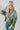 Frontal view of the Leave The Pieces Top that features a green silk material, a multi-colored floral print, a collar neck, a button-up front, long cuffed sleeves, a curved bottom hem, and an oversized fit.