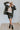 Front view of model wearing the Don't Blame Me Fringe Shacket, that is black faux-leather with a collar, a button-up front, long cuffed sleeves, fringe detailing at the chest, side pockets, and a curved hem. Worn over white tee.