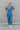 Full body front view of model wearing the Catch Up Denim Jumpsuit, that features a blue colored denim material, a collar and lapel, a surplice neckline, a short cuffed sleeve, front pockets, a button and zipper closure, side pockets, and a straight leg fi
