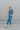 Full body side view of model wearing the Catch Up Denim Jumpsuit, that features a blue colored denim material, a collar and lapel, a surplice neckline, a short cuffed sleeve, front pockets, a button and zipper closure, side pockets, and a straight leg fit