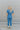 Full body back view of model wearing the Catch Up Denim Jumpsuit, that features a blue colored denim material, a collar and lapel, a surplice neckline, a short cuffed sleeve, front pockets, a button and zipper closure, side pockets, and a straight leg fit