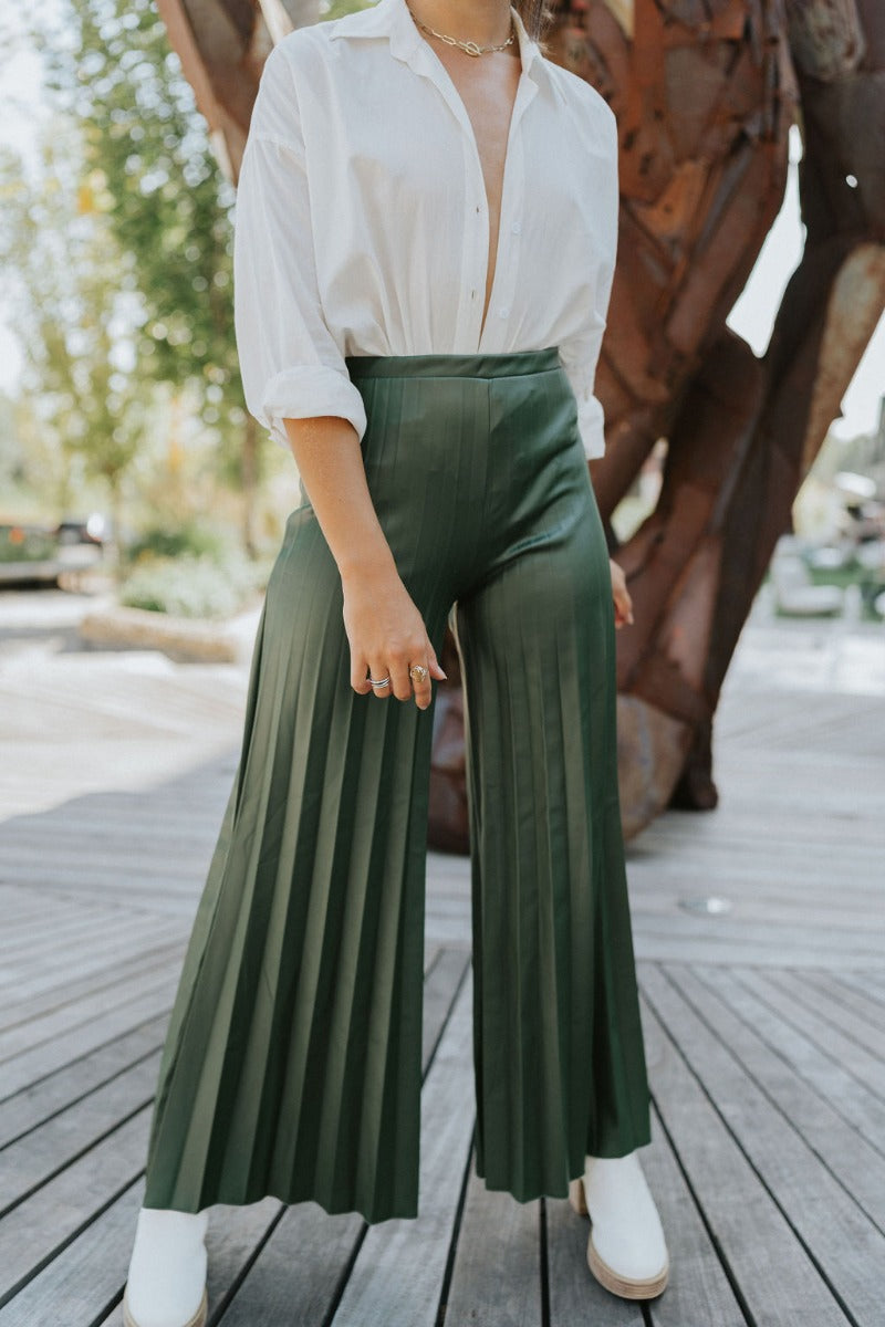 Front view of model wearing the Make You Mine Pants, which feature a hunter green faux-leather material, a pleated design, an elastic waist, and a wide leg fit. Worn with white top.