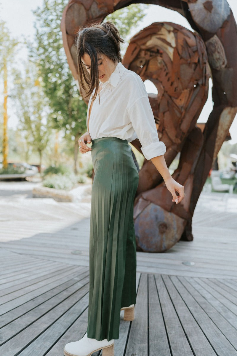 Full body side view of model wearing the Make You Mine Pants, which feature a hunter green faux-leather material, a pleated design, an elastic waist, and a wide leg fit. Worn with white top.