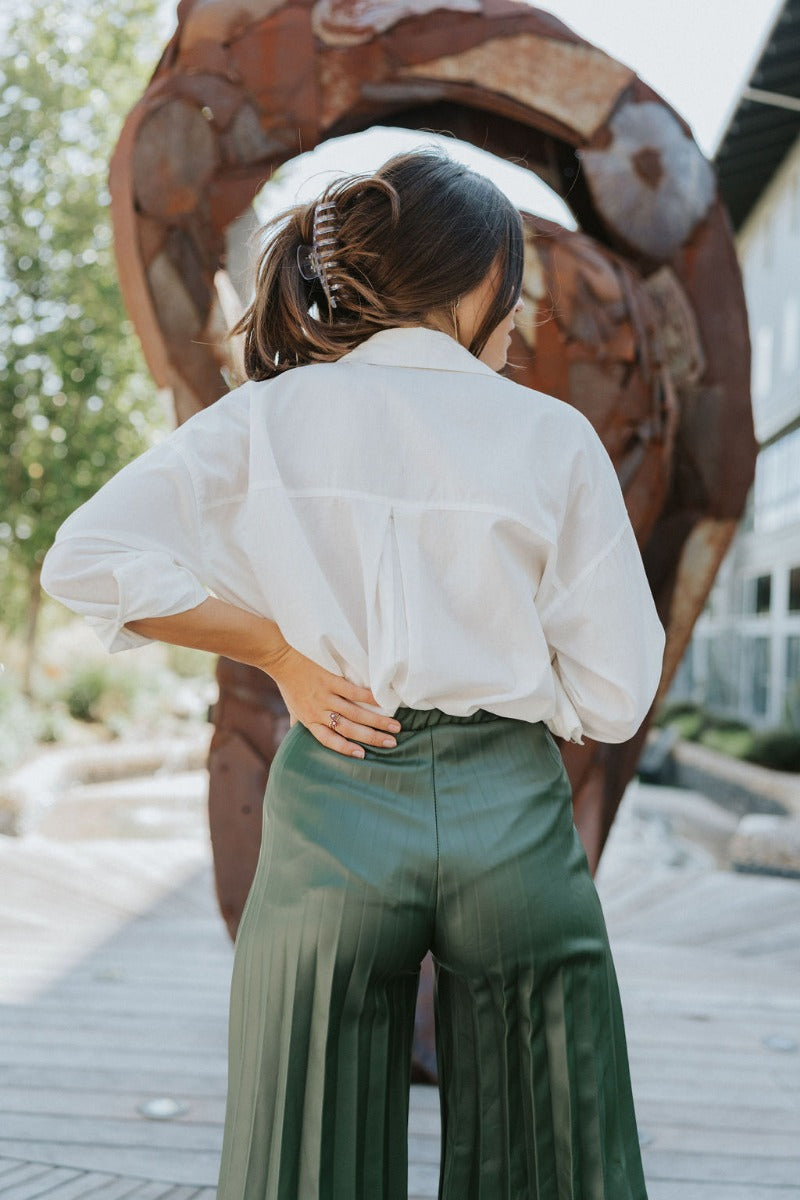 Close-up top/back view of model wearing the Make You Mine Pants, which feature a hunter green faux-leather material, a pleated design, an elastic waist, and a wide leg fit. Worn with white top.