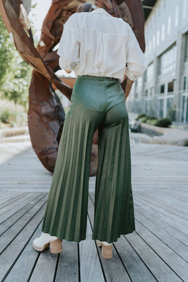 Back view of model wearing the Make You Mine Pants, which feature a hunter green faux-leather material, a pleated design, an elastic waist, and a wide leg fit. Worn with white top.